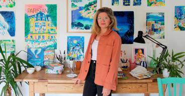 photo d'une jeune femme blonde à lunettes debout avec une veste orange devant un bureau avec du matériel de dessin dessus et devant un mur rempli d'affiches et de dessin