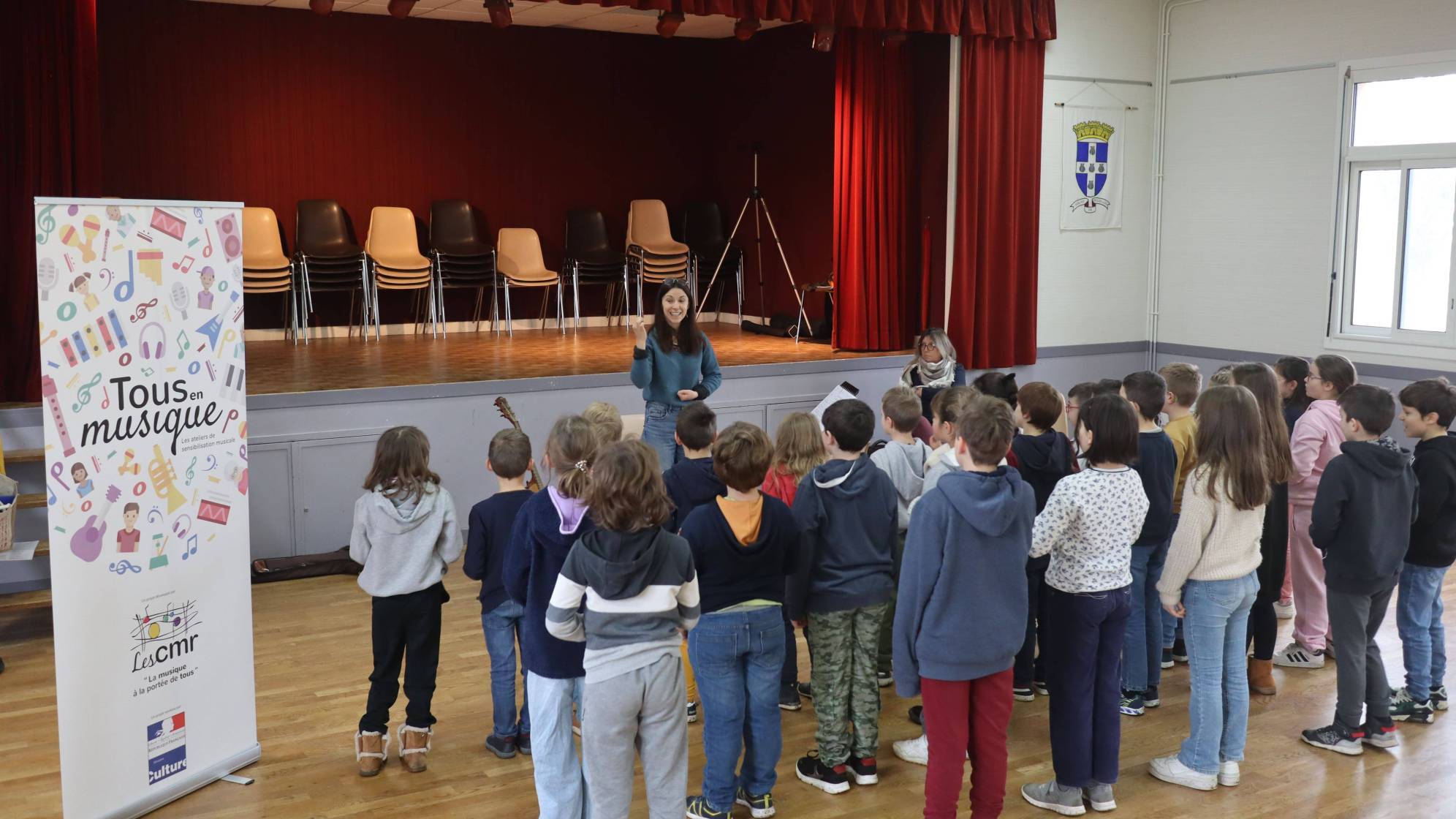 photo d'enfants dans une salle de répétition, de dos, debout en train de chanter avec une professeur