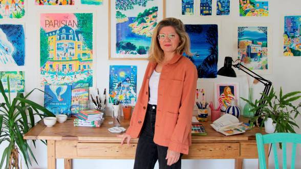 photo d'une jeune femme blonde à lunettes debout avec une veste orange devant un bureau avec du matériel de dessin dessus et devant un mur rempli d'affiches et de dessin